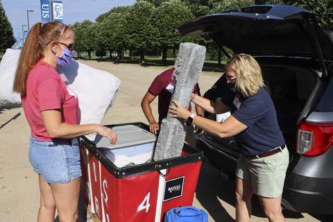 Volunteers unload bedding and supplies from a car into a red move-in cart outside a residence hall.