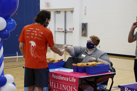 Residence Life staff member hands materials to a student at a check-in table inside a campus gym.
