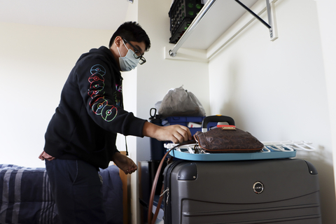 Student wearing a mask unpacks items from a suitcase inside a residence hall room.
