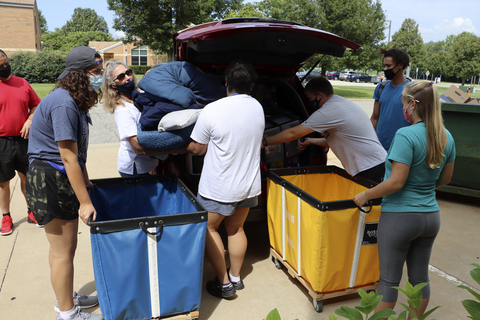 Students and volunteers unload bedding from a car into large move-in carts outside a residence hall.