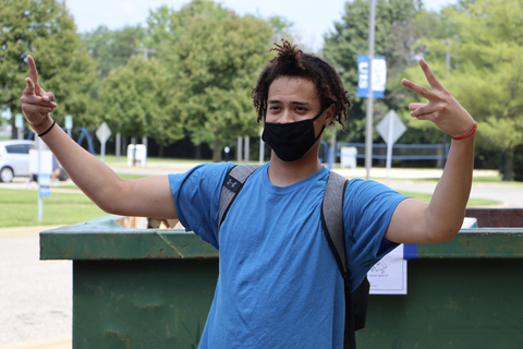 Student wearing a mask smiles and flashes peace signs while standing near move-in carts on campus.