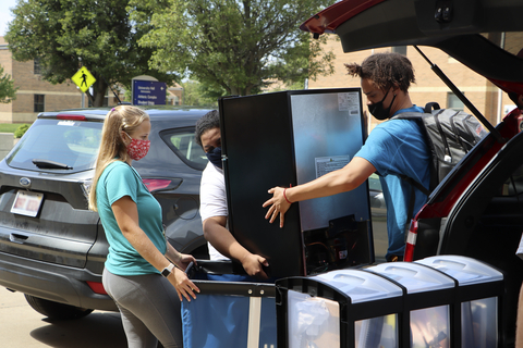 Students and volunteers lift a TV from a car into a blue residence hall move-in cart.