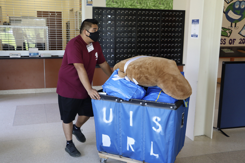 Student pushes a blue UIS residence life cart filled with bedding and belongings inside a residence hall.